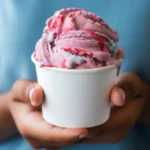 A person in a blue shirt holding a white paper cup filled with scoops of pink and purple swirled ice cream.