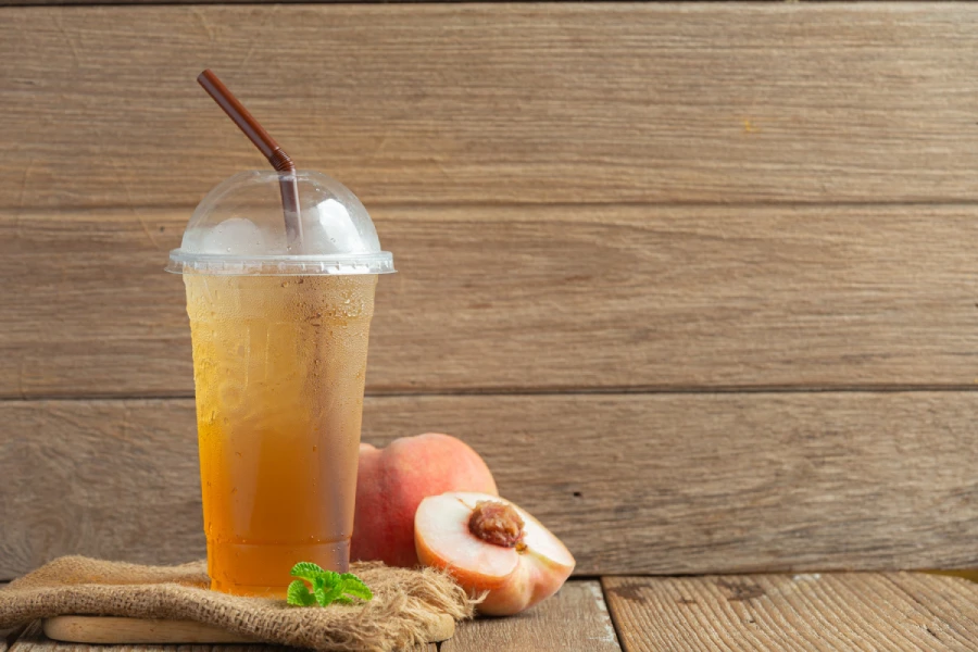 Iced peach tea in a clear plastic cup with a dome lid and brown straw, placed next to a fresh peach on a wooden table.