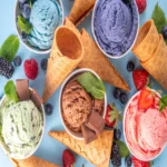 Assorted colorful ice cream scoops in bowls with waffle cones, fresh berries, mint leaves, and chocolate on a pastel blue background.