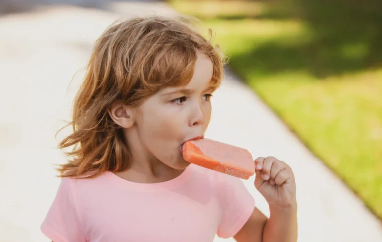 Young child in a pink shirt enjoying a red popsicle outdoors on a sunny day.