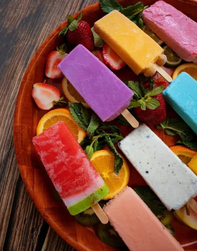 Assorted colorful fruit popsicles on a wooden platter with slices of orange, kiwi, strawberries, and mint leaves.