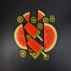 Two red and green popsicles surrounded by watermelon slices and kiwi rounds on a dark background.