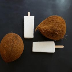 Two white coconut popsicles placed beside whole coconuts on a dark background.