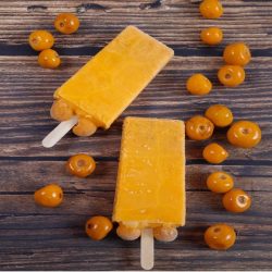 Two yellow popsicles with nance fruits embedded at the base, surrounded by fresh nance on a rustic wooden surface.