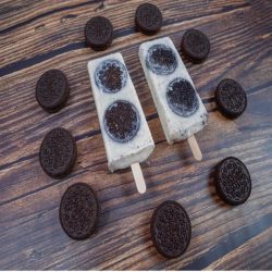 Two cookies and cream popsicles with whole chocolate sandwich cookies, surrounded by cookies on a rustic wooden surface.